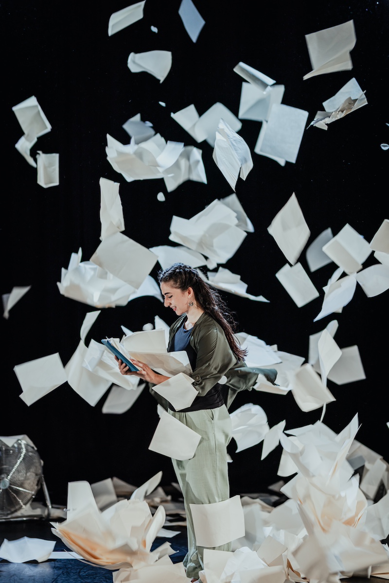 a performer stands reading in the middle of flying sheets of paper