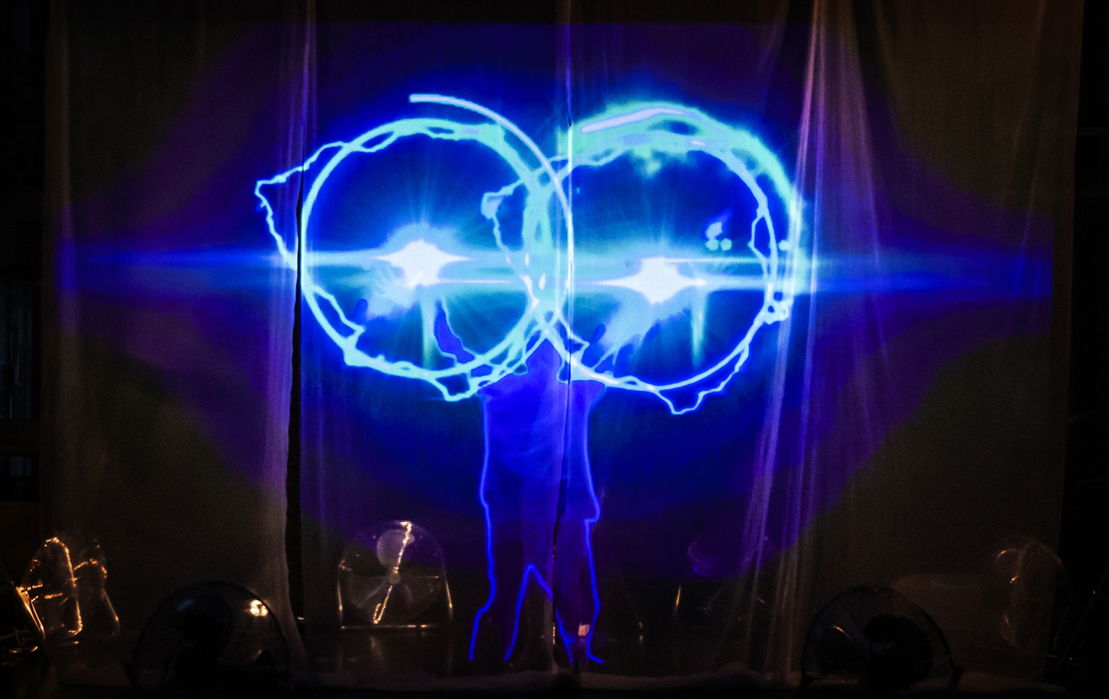 a photo from the performance, with Aakash standing behind a screen, projecting two rings of blue electric light from his outstretched hands