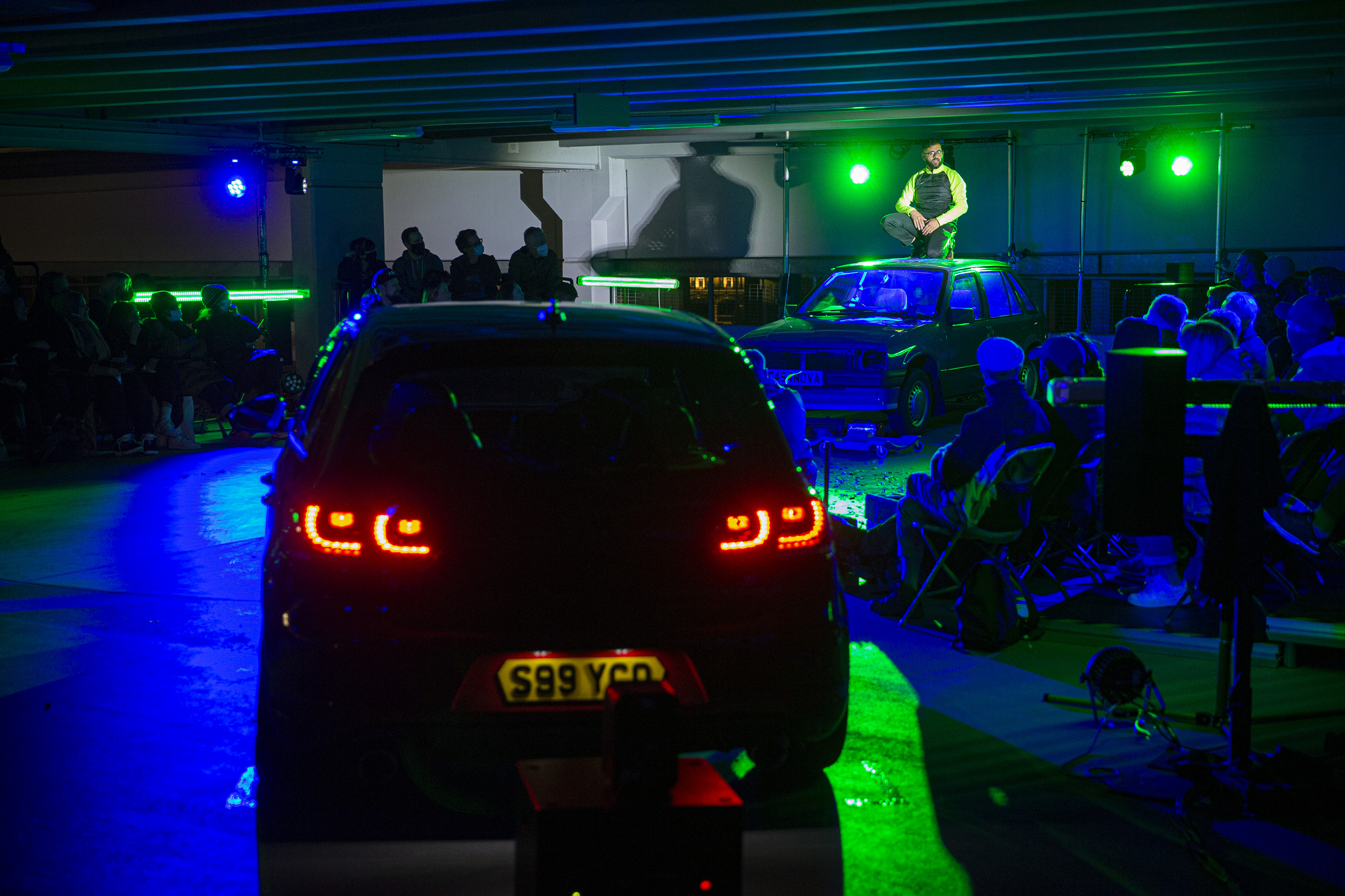 modified cars lit by dramatic lighting in a car park, with an audience watching