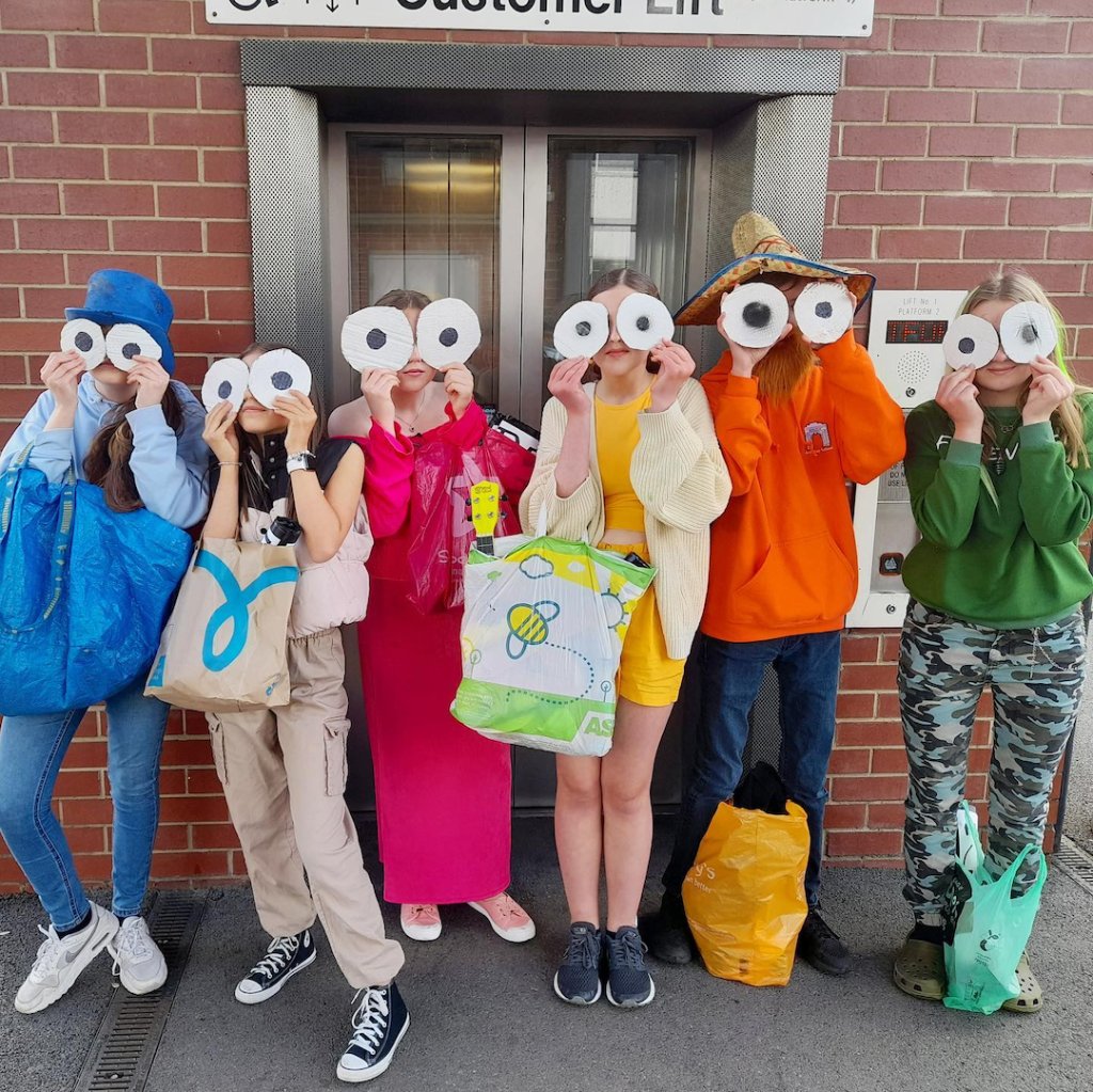 members of Strike A Light Youth Theatre gathered at a rail station holding googly eyes over their faces before a performance on a train