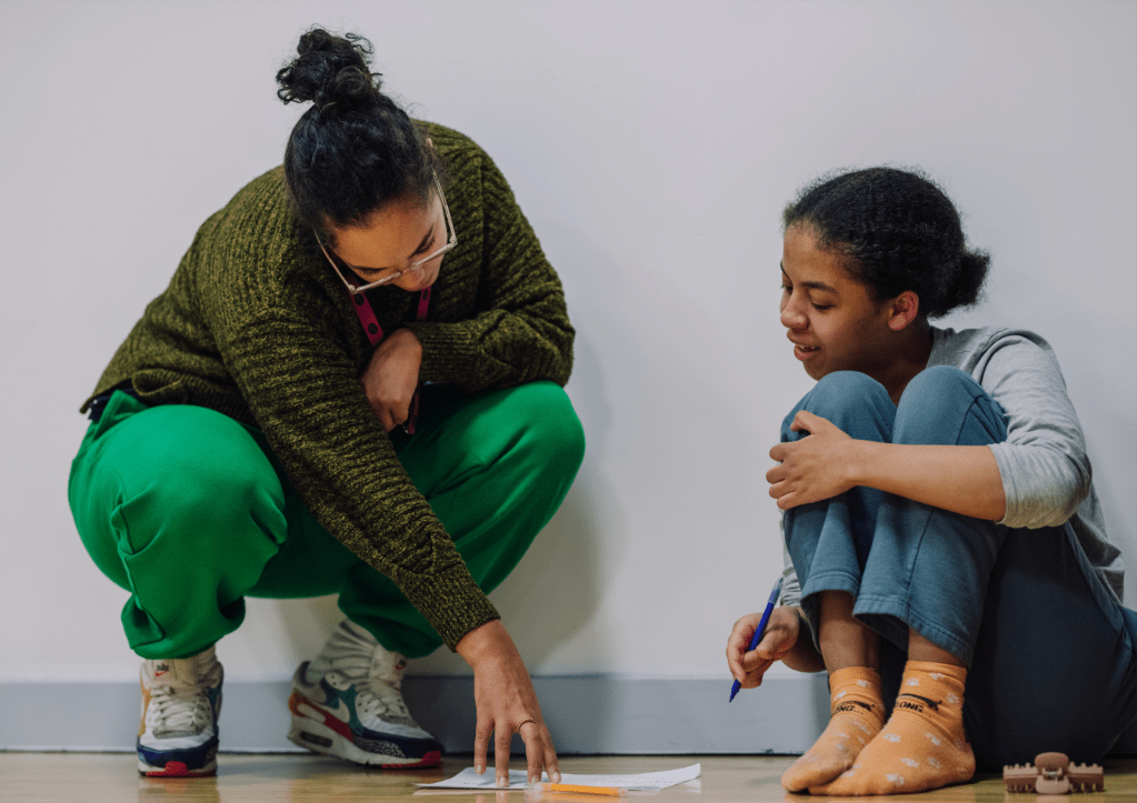 Two women squatting on the floor with a piece of paper between them. One readers the page while holding it down, the other looks on with a pen in hand