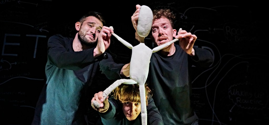 Fred, a two-foot-tall white cloth puppet, is stood dramatically with his arms and one leg stuck out, lit up against a darker background. His three puppeteers, who control his different limbs, are wearing black and looking expressive as they move Fred into position. 